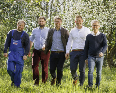 KI generiert: Fünf Männer gehen lächelnd über eine grüne Wiese vor einer Kulisse von blühenden Bäumen. Sie tragen unterschiedliche Kleidung, von Arbeitskleidung bis zu Freizeitkleidung.