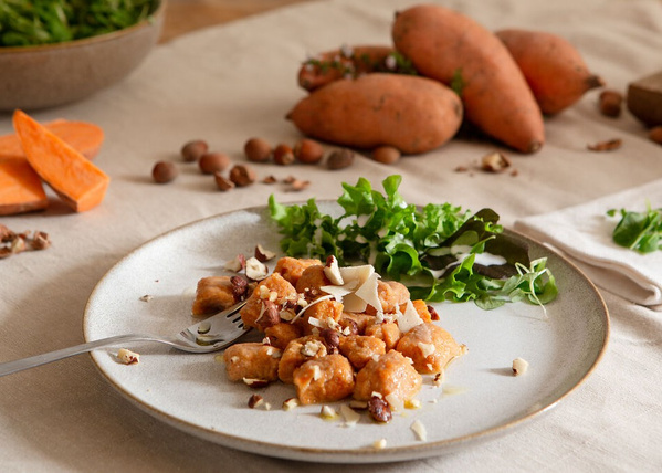 KI generiert: Ein Teller mit Süßkartoffelgnocchi und Salat, im Hintergrund Süßkartoffeln und Haselnüsse.