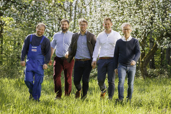 KI generiert: Fünf Männer gehen lächelnd über eine grüne Wiese vor einer Kulisse von blühenden Bäumen. Sie tragen unterschiedliche Kleidung, von Arbeitskleidung bis zu Freizeitkleidung.