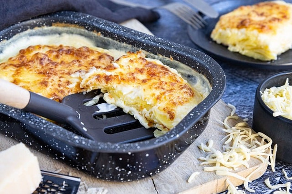 KI generiert: Das Bild zeigt ein frisch gebackenes Kartoffelgratin in einer Auflaufform, aus dem ein Stück herausgenommen wurde. Daneben liegt etwas geriebener Käse als Zutat.