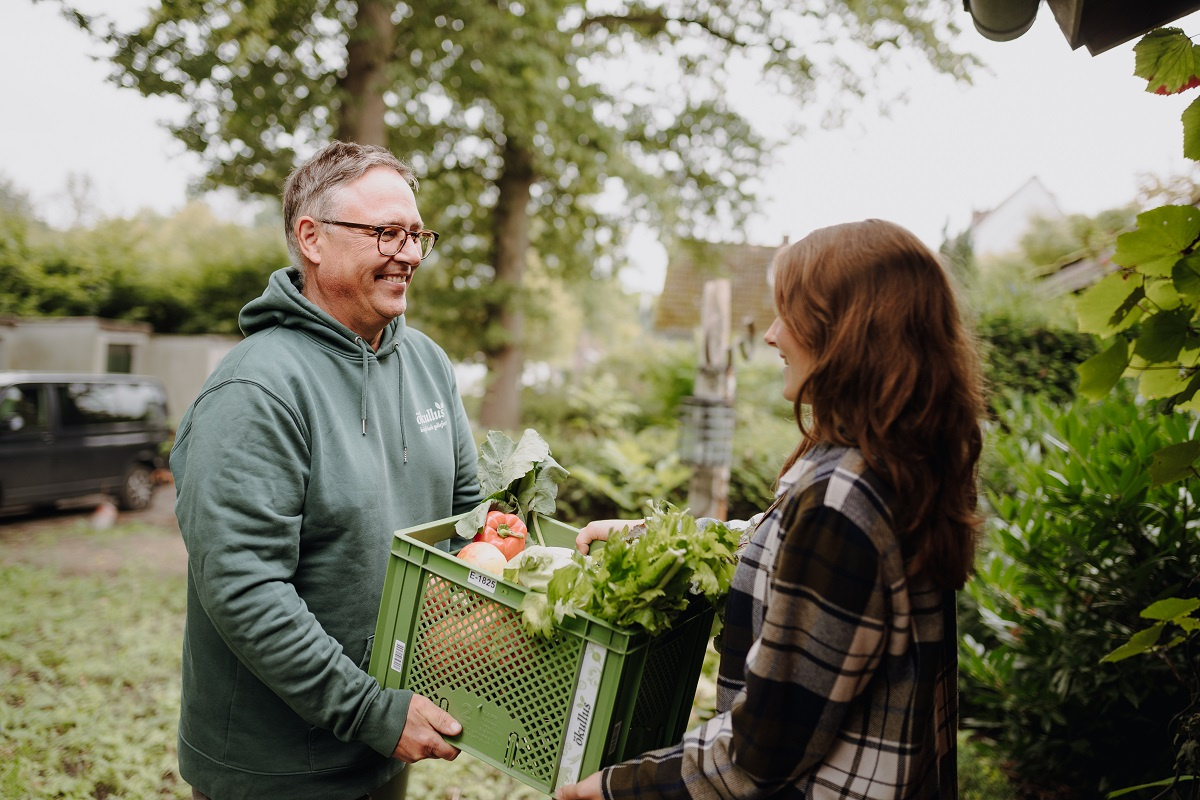 KI generiert: Ein Mann übergibt einer Frau eine grüne Kiste mit frischem Gemüse.