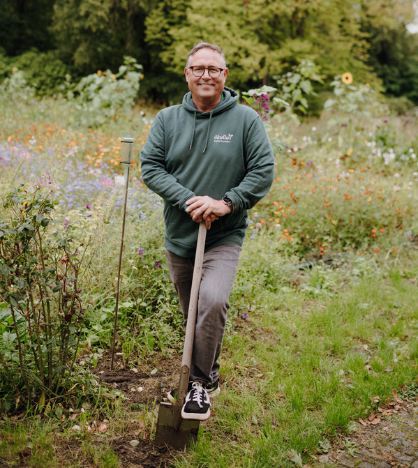 KI generiert: Eine Person lehnt auf einer Schaufel in einem blühenden Garten.