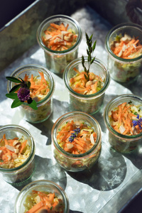 KI generiert: Das Bild zeigt mehrere Gläser, die mit einem bunten Salat aus Karotten, Nüssen und Kräutern gefüllt sind. Jedes Glas ist kunstvoll mit Blüten und Pflanzendekorationen garniert.