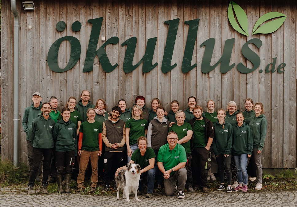 KI generiert: Eine Gruppe von Menschen steht vor einem Holzgebäude mit dem Text "ökullus.de".
