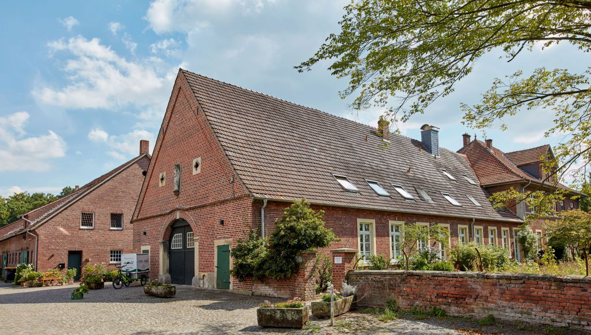 KI generiert: Ein großer, traditioneller Backsteinbauernhof mit begrüntem Innenhof und klarem Himmel.