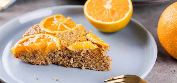 KI generiert: Das Bild zeigt ein Stück Kuchen mit Orangenscheiben auf einem Teller. Daneben liegt eine halbe und eine ganze Orange.