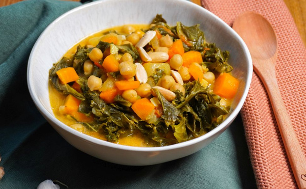 KI generiert: Das Bild zeigt eine Schüssel mit einem bunten Eintopf aus Kichererbsen, Kürbis und grünem Blattgemüse. Daneben liegen eine Stoffserviette und ein Holzlöffel.