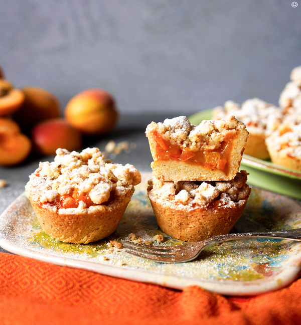 KI generiert: Aprikosen-Streuselmuffins auf einem Teller, einer zerschnitten; im Hintergrund frische Aprikosen.