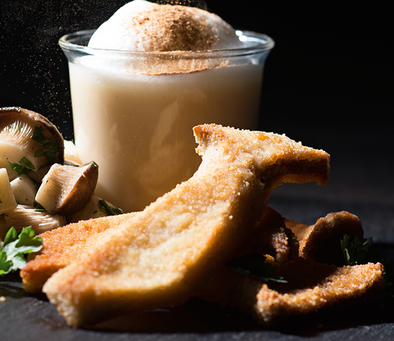 KI generiert: Das Bild zeigt ein elegantes Essen mit panierten Pilzen, gekochten Pilzen und einer Art Soße oder Suppe in einem Glas. Die Präsentation auf einer dunklen Platte lässt ein anspruchsvolles Gericht vermuten.