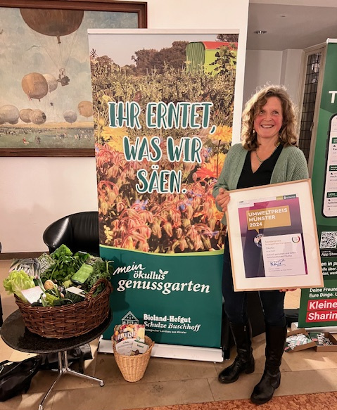 KI generiert: Eine Frau steht neben einem Banner und hält eine Auszeichnung mit der Aufschrift "Umweltpreis Münster 2023" in der Hand. Neben ihr befindet sich ein Korb mit Gemüse.
