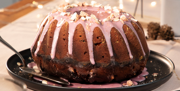 KI generiert: Das Bild zeigt einen Gugelhupf mit rosa Glasur auf einem schwarzen Teller, verziert mit gehackten Nüssen. Der Kuchen ist auf einem gedeckten Tisch angerichtet.