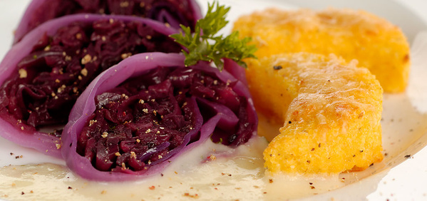 KI generiert: Das Bild zeigt ein Gericht mit Rotkohlscheiben, die gefüllt zu sein scheinen, sowie goldbraun gebratene Polentaschnitten auf einer weißen Sauce. Eine kleine Garnitur aus Kräutern rundet das Arrangement ab.