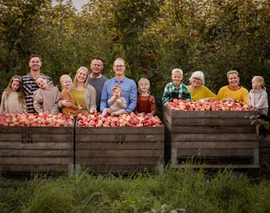 KI generiert: Das Bild zeigt eine Gruppe von Menschen, die hinter großen Kisten voller Äpfel im Freien posieren. Sie stehen auf einer Wiese, umgeben von Apfelbäumen.