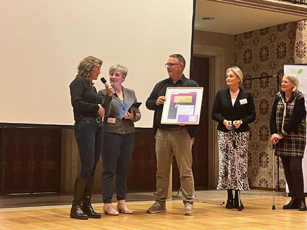 KI generiert: Das Bild zeigt eine Gruppe von fünf Menschen auf einer Bühne, wobei eine Person eine Auszeichnung entgegennimmt. Zwei Frauen haben Mikrofone in der Hand und sprechen zu den anderen.