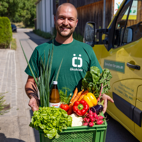 KI generiert: Ein Mann in einem grünen Shirt mit dem Logo "ökokiste" hält eine Kiste voller frischem Gemüse und einer Flasche Saft vor einem gelben Lieferwagen. Der Hauptinhalt des Bildes ist die Präsentation von frischen Bioprodukten.