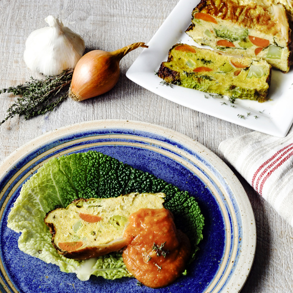 KI generiert: Das Bild zeigt ein Stück Gemüsestrudel auf einem grünen Salatblatt, garniert mit einer Tomatensoße auf einem blauen Teller. Im Hintergrund befinden sich Knoblauch, eine Zwiebel und frische Kräuter zur Dekoration.