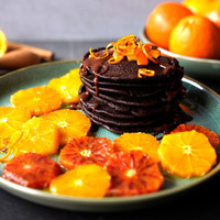 KI generiert: Das Bild zeigt einen Teller mit einem Stapel dunkler Schokoladenpfannkuchen, garniert mit Orangenscheiben und Schokoladensauce. Im Hintergrund sind frische Orangen zu sehen.