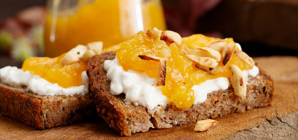 KI generiert: Das Bild zeigt zwei Scheiben Vollkornbrot, die mit Frischkäse, Mango-Chutney und gehackten Nüssen garniert sind. Sie liegen auf einem hölzernen Untergrund, im Hintergrund ist verschwommen ein Glas mit Chutney zu sehen.