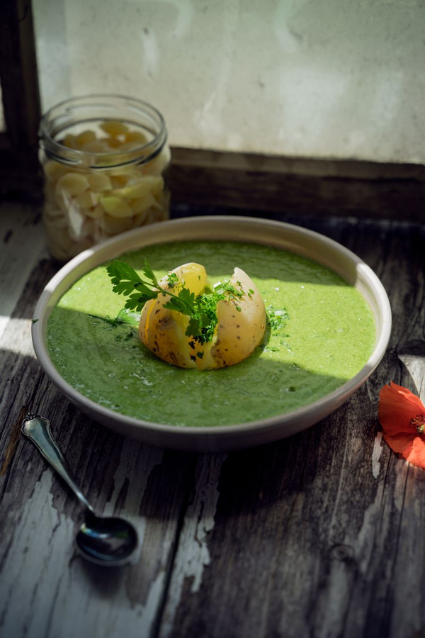 KI generiert: Das Bild zeigt eine Schüssel mit einer grünen Suppe, die mit einer halben Kartoffel und frischen Kräutern garniert ist. Im Hintergrund ist ein Glas mit eingelegtem Gemüse zu sehen.