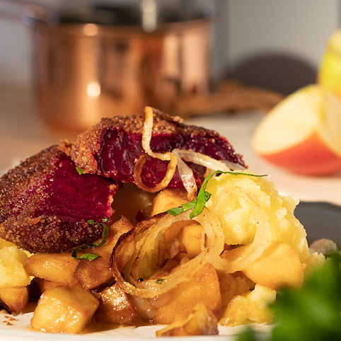 KI generiert: Ein Gericht mit panierten Rote-Bete-Scheiben, Kartoffeln und Zwiebeln auf einem Teller, im Hintergrund ein Topf.