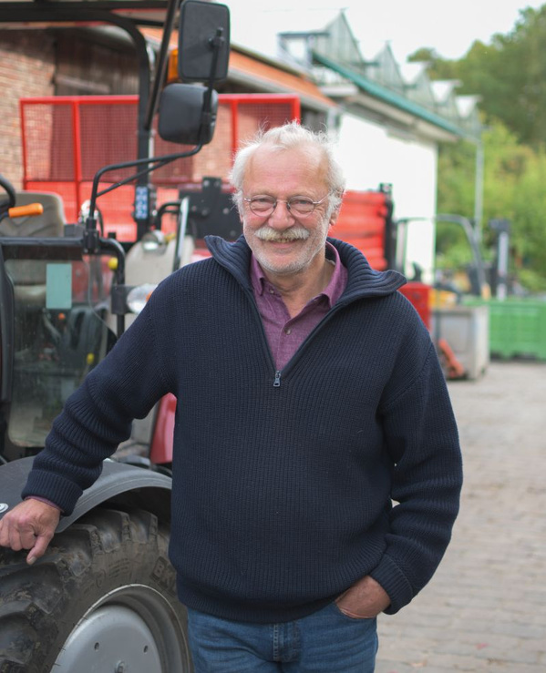 KI generiert: Ein älterer Mann lächelt in die Kamera und steht neben einem Traktor auf einem Hof. Der Hintergrund zeigt landwirtschaftliche Gebäude und Maschinen.