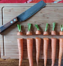 KI generiert: Auf dem Bild sind mehrere ganz und teilweise geschnittene Karotten auf einem Holzschneidebrett zu sehen, zusammen mit einem großen Messer. Die Karotten sind so platziert, dass die oberen Teile gleichmäßig abgetrennt nebeneinander liegen.