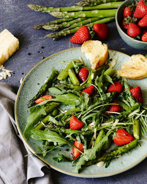 KI generiert: Das Bild zeigt einen frischen Salat mit Spargel, Erdbeeren und Rucola, garniert mit Brotscheiben und Käsestücken. Daneben liegen weitere Zutaten wie Spargel und Erdbeeren auf einem dunklen Untergrund bereit.