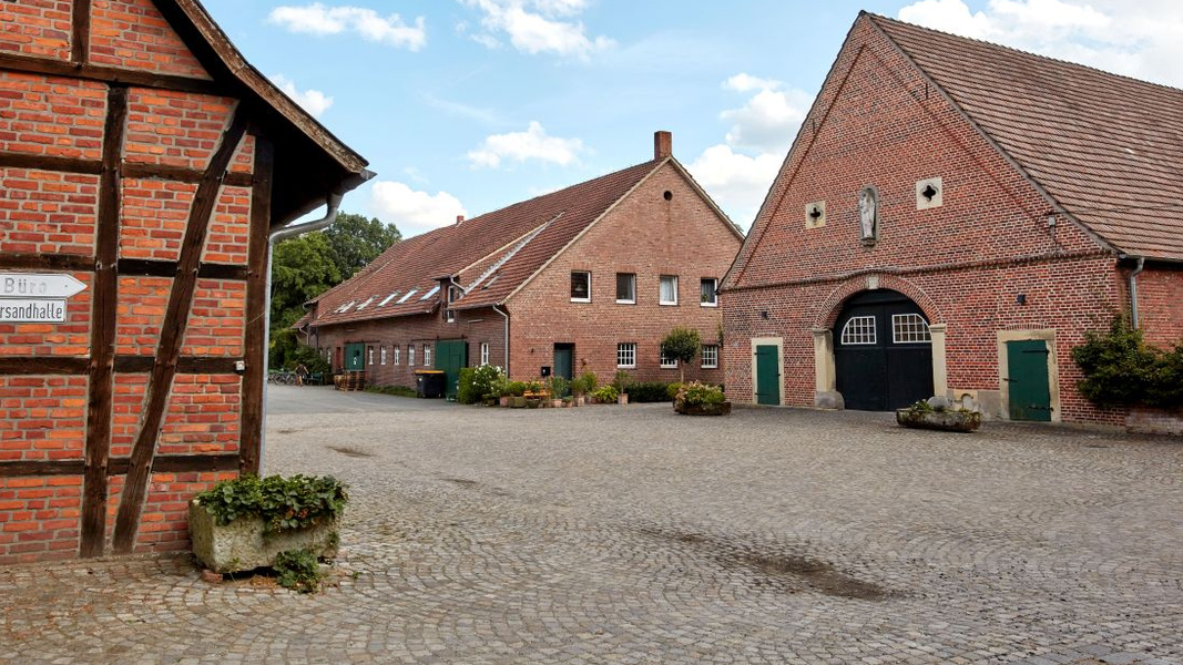 KI generiert: Ein Bauernhof mit roten Ziegelgebäuden. Text: "Büro Versandhalle".