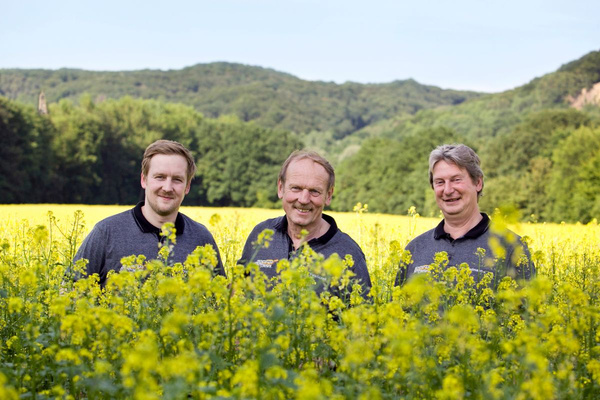 KI generiert: Drei Männer stehen lächelnd in einem blühenden Rapsfeld, umgeben von grünen Hügeln im Hintergrund. Der Hauptinhalt des Bildes ist die ländliche Szenerie mit den Männern im Vordergrund.