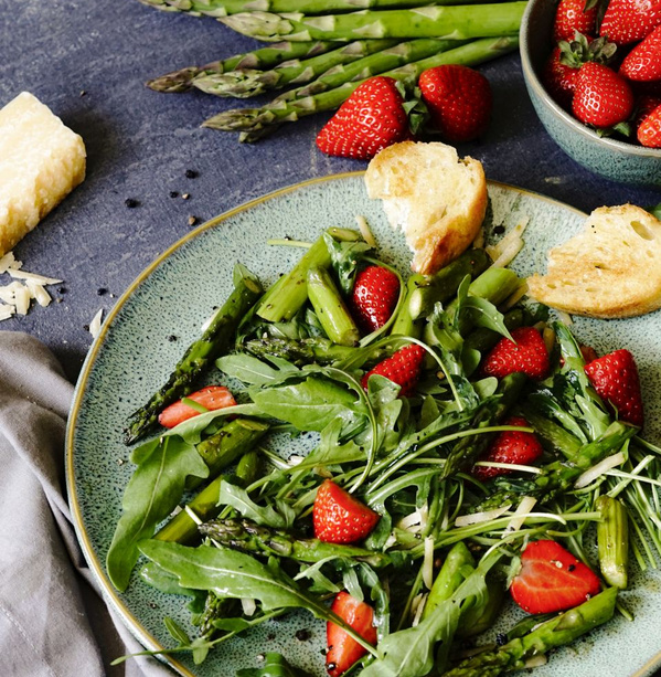 KI generiert: Das Bild zeigt einen frischen Salat mit Spargel, Erdbeeren und Rucola, garniert mit Brotscheiben und Käsestücken. Daneben liegen weitere Zutaten wie Spargel und Erdbeeren auf einem dunklen Untergrund bereit.