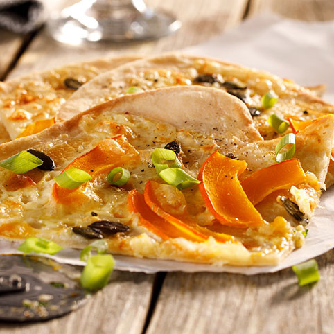 KI generiert: Zwei Stücke Flammkuchen mit Kürbis, Frühlingszwiebeln und Kernen auf Pergamentpapier auf einem Holztisch.