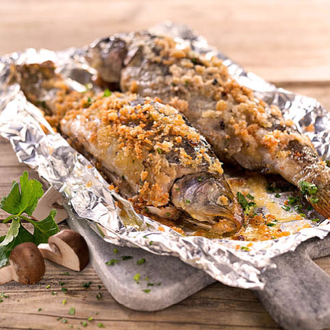 KI generiert: Zwei gegrillte Fische auf Alufolie mit einer Kräuter-Gemüse-Garnitur auf einem Holzbrett.