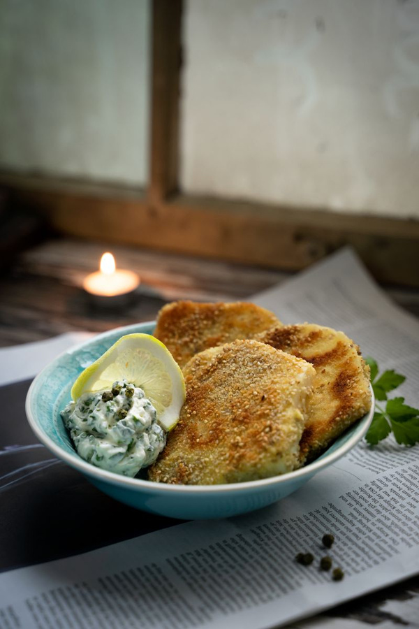 KI generiert: Das Bild zeigt ein Gericht aus paniertem, gebratenem Fisch oder Fleisch, serviert mit einem Zitronenschnitz und einer Kräutercreme, in einer Schüssel auf einer Zeitung. Im Hintergrund brennt eine Kerze und sorgt für stimmungsvolles Licht.