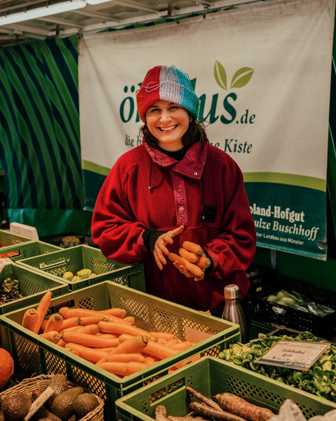 KI generiert: Eine Person steht hinter Gemüsekisten auf einem Marktstand. Text: "oekolus.de - die Heimat der Biokiste".