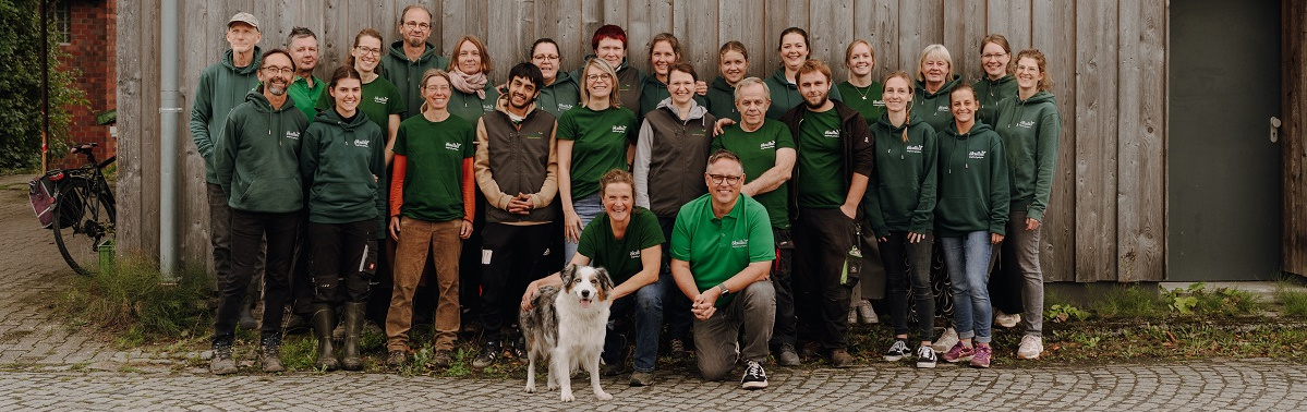 KI generiert: Eine Gruppe von 24 Personen und einem Hund posiert vor einer Holzwand.