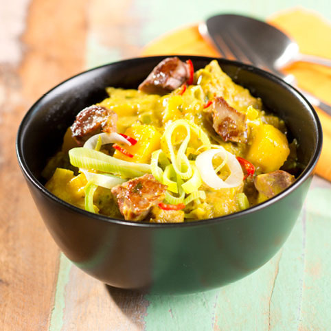 KI generiert: Schwarze Schale mit Currygericht, enthält Fleischstücke, Lauch und Paprika auf hellem Holztisch.