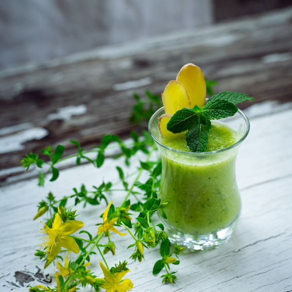 KI generiert: Grüner Smoothie in einem Glas, garniert mit Minze und Ingwer, auf rustikalem Holztisch, umgeben von gelben Blumen.