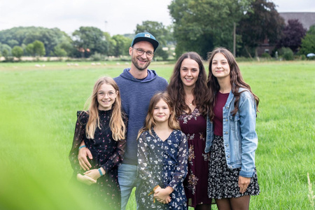 KI generiert: Das Bild zeigt eine Gruppe von fünf Personen, vermutlich eine Familie, die draußen auf einer grünen Wiese steht und lächelt. Im Hintergrund sind Bäume und ein Gebäude zu sehen.