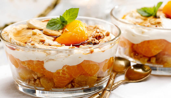 KI generiert: Das Bild zeigt zwei Schalen mit einem Dessert, das aus Schichten von Mandarinen, Sahne und Nüssen besteht und mit einem Minzblatt und karamellisierten Aprikosenstücken garniert ist. Der Hauptinhalt des Bildes ist das dekorativ angerichtete Dessert in einer Glasschale.