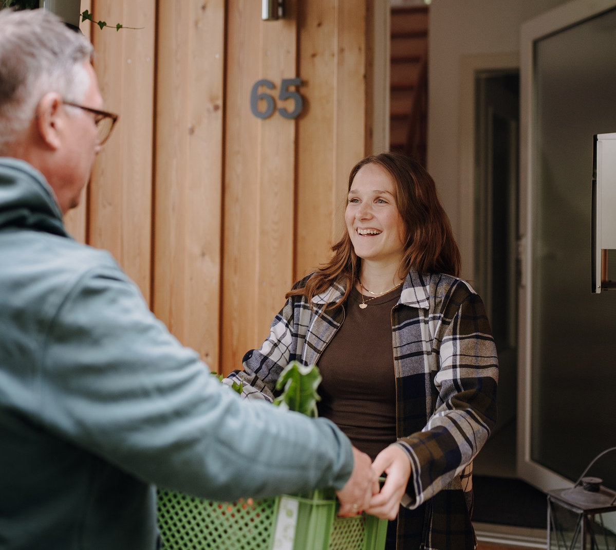 KI generiert: Eine Person nimmt draußen eine Gemüsekiste von jemandem entgegen, lächelnd. Hausnummer 65.