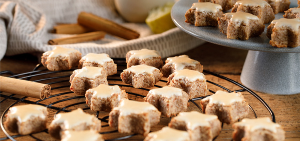KI generiert: Auf dem Bild sind zimtsterne, traditionelle deutsche Plätzchen mit Zuckerguss, auf einem Gitterrost und einem Teller zu sehen. Im Hintergrund befinden sich Zimtstangen und ein Stofftuch.